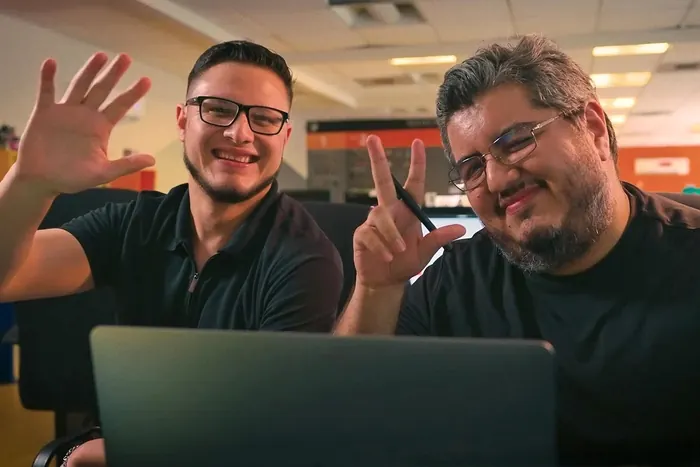 Two CGBOT teammates smiling and waving at the camera from behind a laptop in the studio
